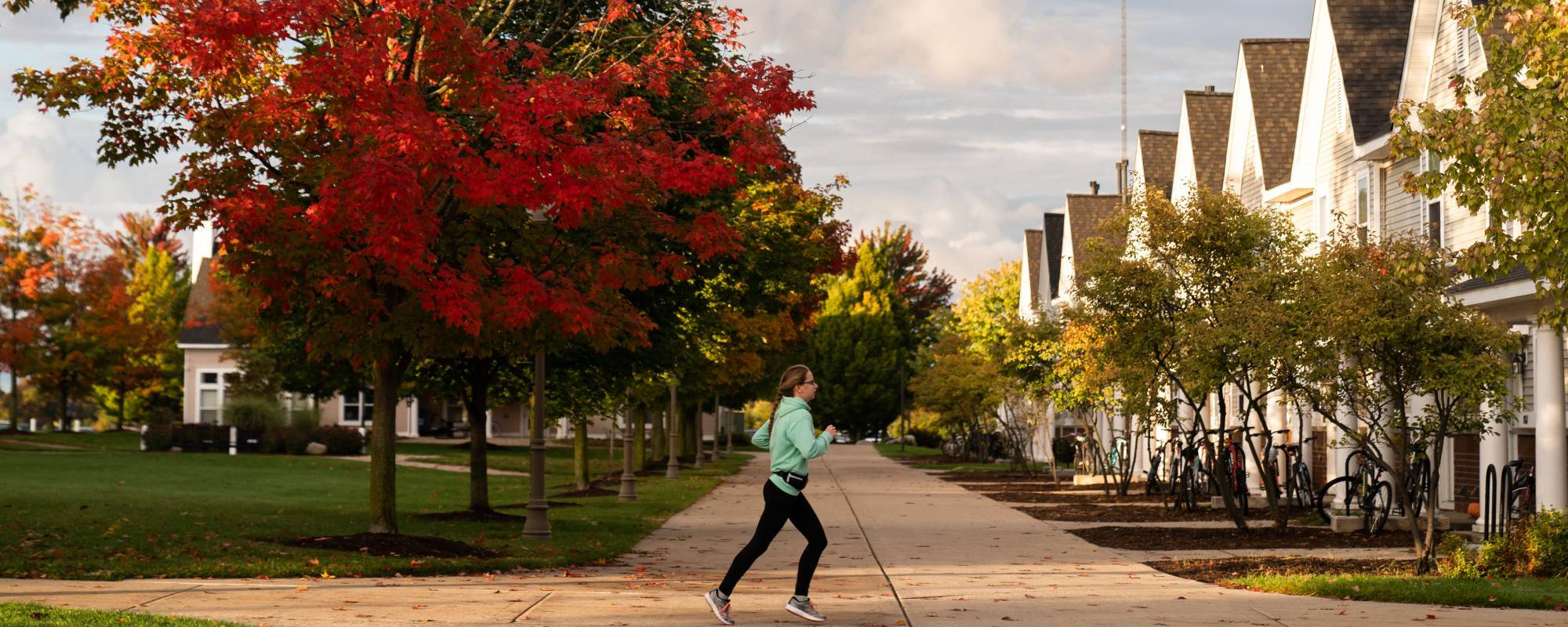 A person jogs on a tree-lined suburban sidewalk outside the Laker Village Apartments under a vibrant autumn tree with red leaves. The street is lined with white houses and bikes.